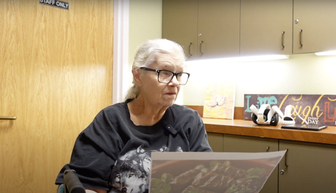 An older woman with light hair tied back and wearing glasses and a black T-shirt featuring wolves sits at a chair, holding a picture of dolmades. Her gaze is thoughtful and nostalgic as she gently interacts with the photo.